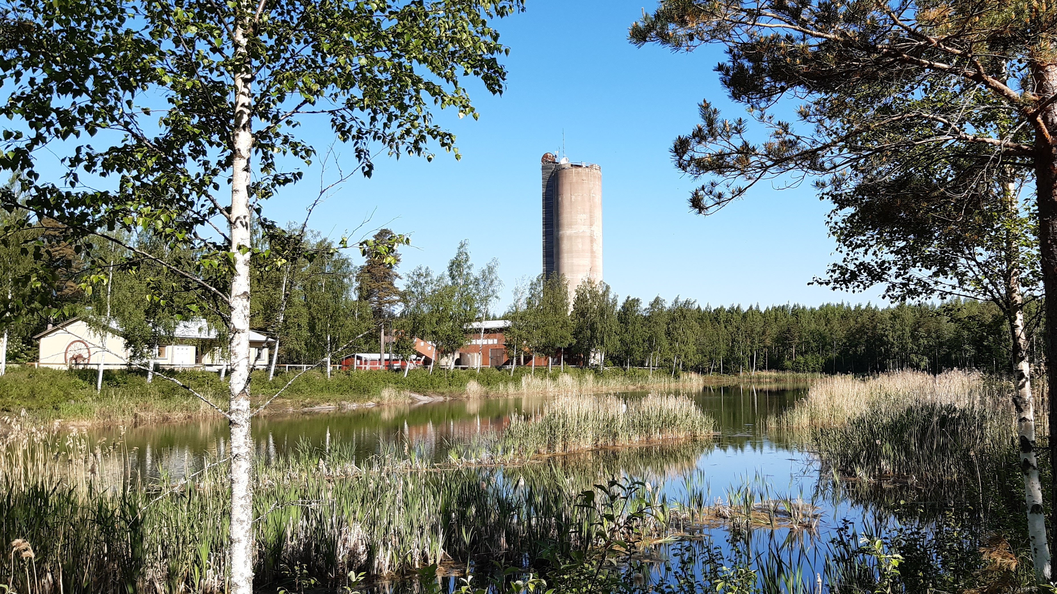 Etualalla puita ja vettä, taaempana pieniä rakennuksia ja kaivostorni.