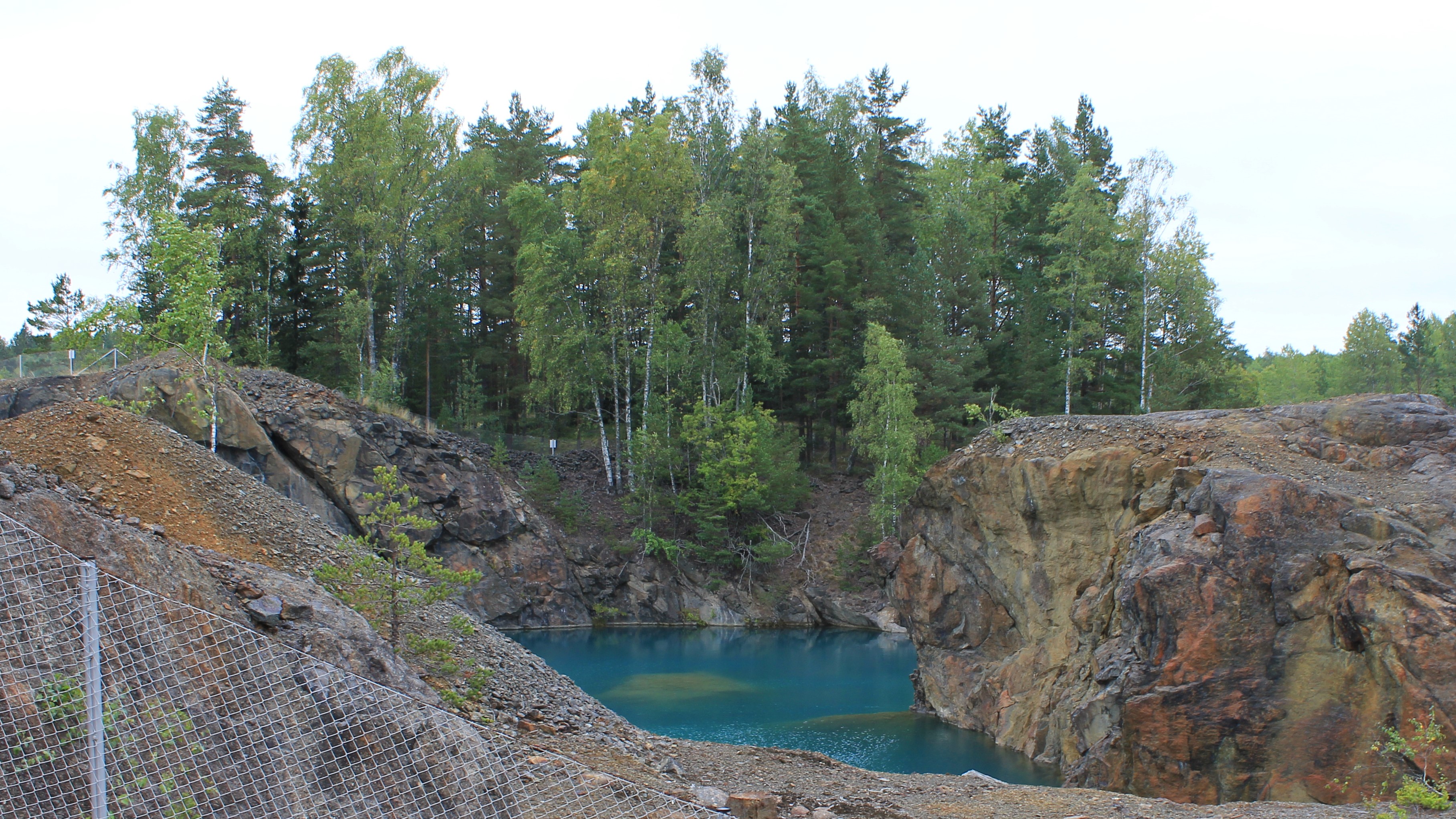 Turkoosilla vedellä täyttynyt avolouhos, jonka ympärillä on kallioita. Taaempana on puita.