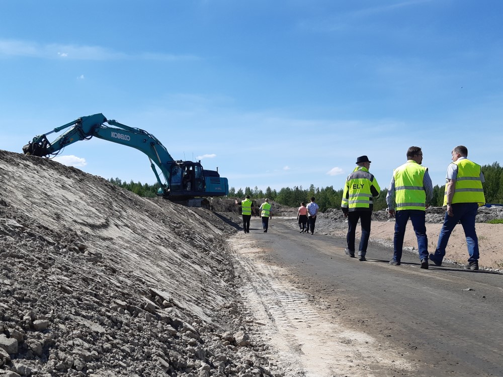 Takana työmaa ja kaivonkone, edempänä huomiotakkisia ihmisiä. Yhden selässä lukee ELY.