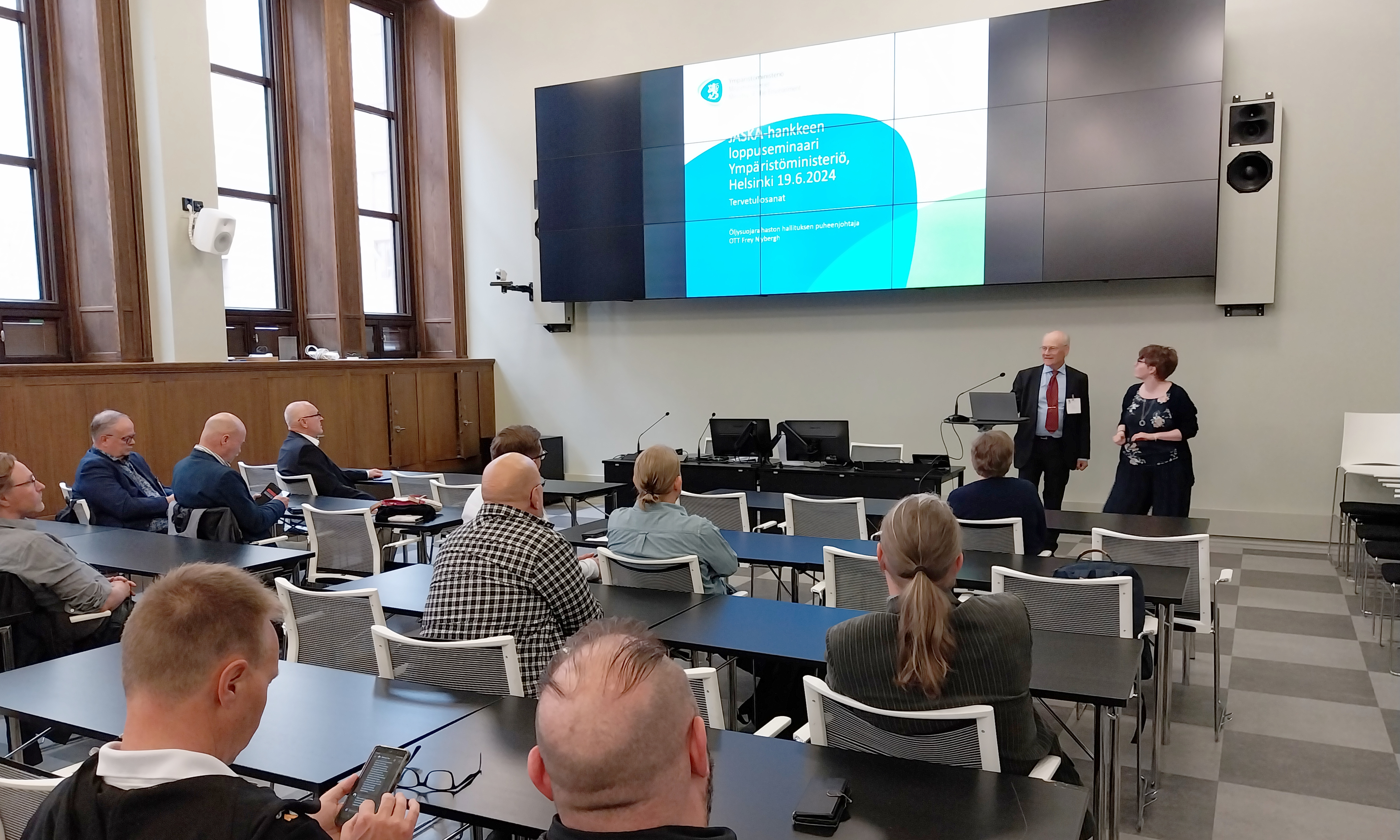 Kuvassa on luentosali ja ihmisiä sekä kaksi puhujaa edessä.