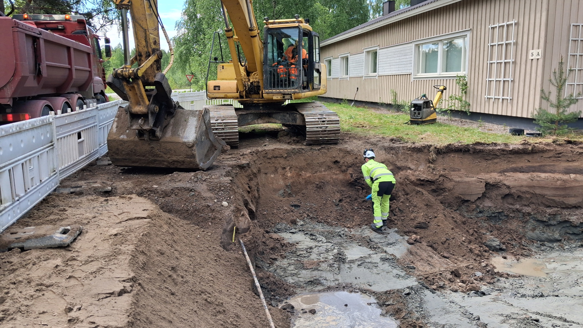 Kuvassa on työmaa ja työmaakone sekä henkilö, joka ottaa maaperänäytettä kaivannon seinämästä.